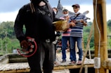 A group of people stand on a bamboo platform, playing musical instruments such as tambourines and guitars. They are casually dressed and appear to be enjoying themselves. The background includes lush greenery and possibly a traditional building with a pointed roof.