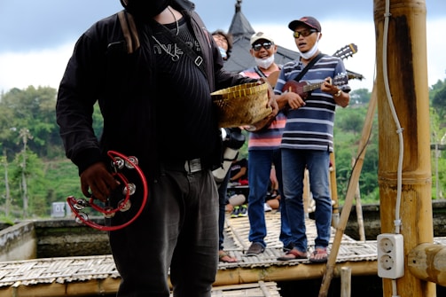 A group of people stand on a bamboo platform, playing musical instruments such as tambourines and guitars. They are casually dressed and appear to be enjoying themselves. The background includes lush greenery and possibly a traditional building with a pointed roof.