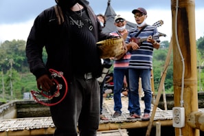 A group of people stand on a bamboo platform, playing musical instruments such as tambourines and guitars. They are casually dressed and appear to be enjoying themselves. The background includes lush greenery and possibly a traditional building with a pointed roof.