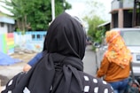 A person wearing a black headscarf with white patterns is in the foreground, facing away from the camera. In the background, another person is walking, wearing a colorful patterned headscarf and a brown jacket. The setting appears to be a residential street with a parked car, trees, and a multicolored painted wall.