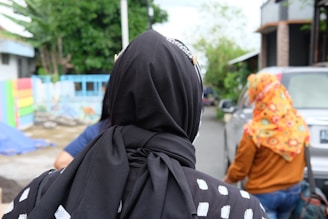 A person wearing a black headscarf with white patterns is in the foreground, facing away from the camera. In the background, another person is walking, wearing a colorful patterned headscarf and a brown jacket. The setting appears to be a residential street with a parked car, trees, and a multicolored painted wall.