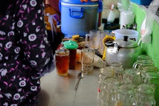 A cluttered kitchen counter with various items, including a metallic kettle, glasses filled with brown liquid, an assortment of utensils, a spoon, and a blue cooler in the background. There are also jars and a plastic container with a green lid, and some ingredients like sugar bags. The scene suggests a busy and casual kitchen environment.