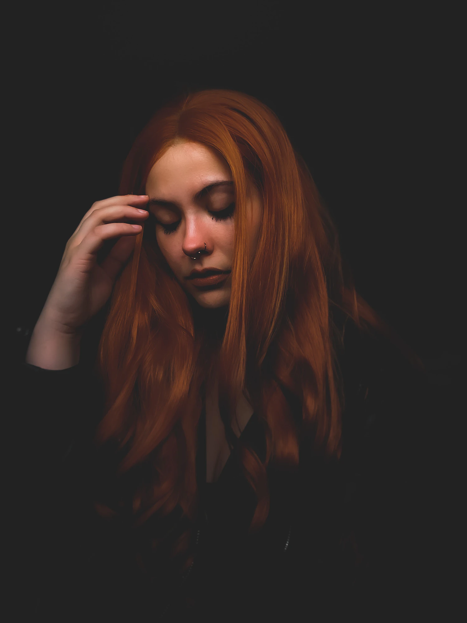 A woman with long red hair in a dark room, one hand lifting a lock away from her temple