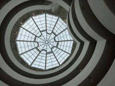 A view upward through a spiraling architectural interior with a glass dome roof. The structure features concentric circles and curves creating a sense of movement and depth.