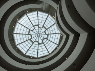 A view upward through a spiraling architectural interior with a glass dome roof. The structure features concentric circles and curves creating a sense of movement and depth.