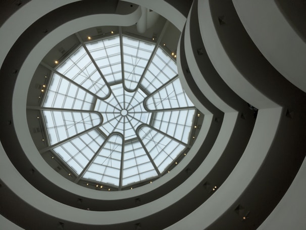 A view upward through a spiraling architectural interior with a glass dome roof. The structure features concentric circles and curves creating a sense of movement and depth.