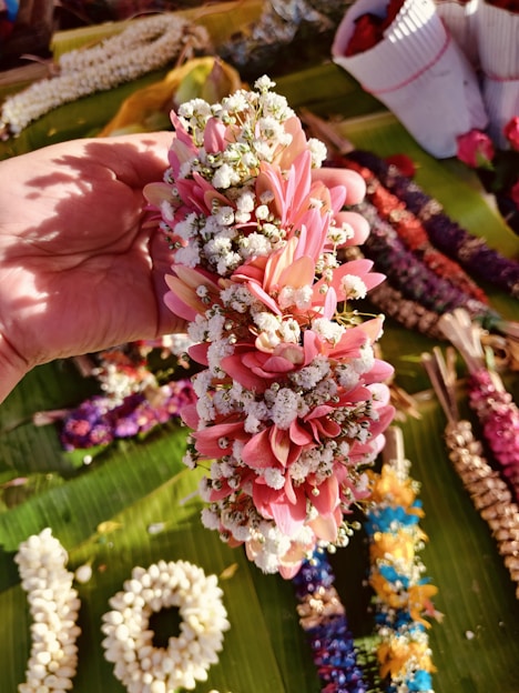 A hand holding a beautiful floral garland made up of small pink and white flowers. The background shows multiple other garlands and flower arrangements placed on a green surface, possibly leaves. There are various colored garlands laid out including purple, yellow, and more.