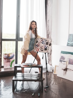 A young woman stands in an art studio, beside a wooden easel holding a canvas with colorful handprints. She wears a light jacket over denim overalls and short denim shorts. The studio has large windows allowing natural light to pour in, illuminating several abstract paintings leaning against the walls.