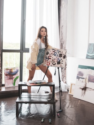 A young woman stands in an art studio, beside a wooden easel holding a canvas with colorful handprints. She wears a light jacket over denim overalls and short denim shorts. The studio has large windows allowing natural light to pour in, illuminating several abstract paintings leaning against the walls.