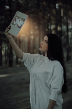a woman holding up a book in a forest and light shining down out of the book's open pages
