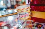 Ice cream cups with the label Delicious Ice Cream are stacked on a reflective surface. The background is a mix of colorful and blurred lights, giving a festive or carnival-like atmosphere. There is a red object to the right, possibly part of a vending machine or a counter, with the number 25 displayed digitally.