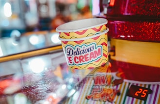 Ice cream cups with the label Delicious Ice Cream are stacked on a reflective surface. The background is a mix of colorful and blurred lights, giving a festive or carnival-like atmosphere. There is a red object to the right, possibly part of a vending machine or a counter, with the number 25 displayed digitally.