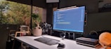 A modern workspace is set up with a large computer monitor displaying code. The desk features a sleek keyboard, mouse, and various tech gadgets. On the desk, there is a potted plant, a stack of closed notebooks, and a spherical lamp emitting warm light. The area receives natural light from a large window, outside of which trees are visible. A model car is seen in the background.