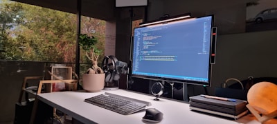 A modern workspace is set up with a large computer monitor displaying code. The desk features a sleek keyboard, mouse, and various tech gadgets. On the desk, there is a potted plant, a stack of closed notebooks, and a spherical lamp emitting warm light. The area receives natural light from a large window, outside of which trees are visible. A model car is seen in the background.