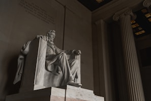 A large marble statue of a seated figure is situated within a grand interior featuring tall columns and an inscribed wall. The statue is intricately carved, capturing the details of the figure's expression and clothing. The lighting is soft, casting subtle shadows that enhance the sculpture's features.