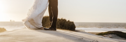 A couple stands on a sandy beach, with the bride's flowing white dress and the groom's formal black suit contrasting against the soft golden light of an approaching sunset. The expansive ocean is visible in the background, with gentle waves meeting the shore under a serene sky.
