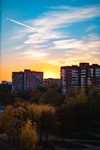 A sunset skyline featuring tall, red brick apartment buildings with many windows, set against a vibrant sky. The sky is painted with a blend of blue, orange, and yellow hues with scattered clouds, and a visible contrail stretching across. In the foreground, there are lush trees with autumn-colored leaves in shades of green, yellow, and orange.