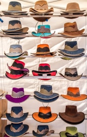 Multiple cowboy hats of various colors and styles are neatly arranged on shelves, each featuring unique designs and details including different textures and band patterns.