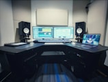 A music production studio setup featuring two computer monitors displaying a digital audio workstation, a laptop with an animated image on the screen, two studio speakers on stands, a MIDI keyboard, and other audio equipment on a curved desk.