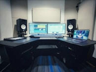 A music production studio setup featuring two computer monitors displaying a digital audio workstation, a laptop with an animated image on the screen, two studio speakers on stands, a MIDI keyboard, and other audio equipment on a curved desk.