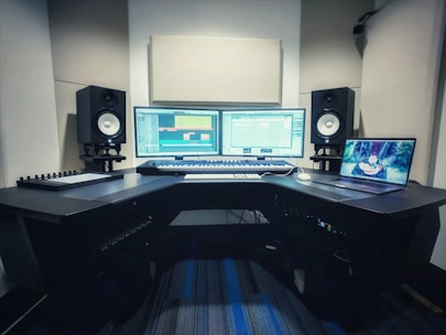 A music production studio setup featuring two computer monitors displaying a digital audio workstation, a laptop with an animated image on the screen, two studio speakers on stands, a MIDI keyboard, and other audio equipment on a curved desk.