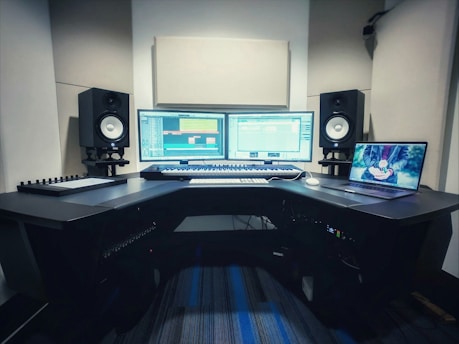 A music production studio setup featuring two computer monitors displaying a digital audio workstation, a laptop with an animated image on the screen, two studio speakers on stands, a MIDI keyboard, and other audio equipment on a curved desk.