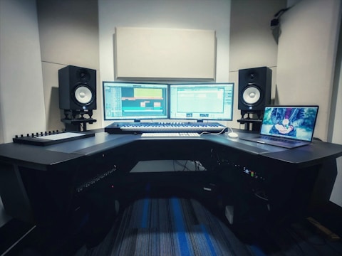 A music production studio setup featuring two computer monitors displaying a digital audio workstation, a laptop with an animated image on the screen, two studio speakers on stands, a MIDI keyboard, and other audio equipment on a curved desk.