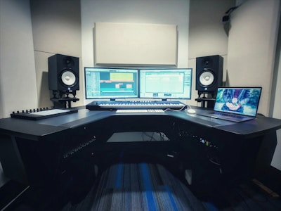 A music production studio setup featuring two computer monitors displaying a digital audio workstation, a laptop with an animated image on the screen, two studio speakers on stands, a MIDI keyboard, and other audio equipment on a curved desk.