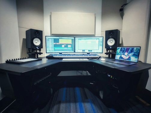 A music production studio setup featuring two computer monitors displaying a digital audio workstation, a laptop with an animated image on the screen, two studio speakers on stands, a MIDI keyboard, and other audio equipment on a curved desk.