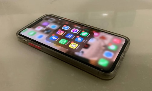 A smartphone lies on a flat surface, displaying several social media app icons including Facebook, Instagram, Twitter, LinkedIn, WhatsApp, Snapchat, and Messenger. The phone is encased in a transparent protective case.