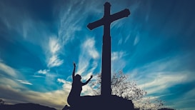 A silhouetted figure is kneeling with raised arms in front of a large cross against a dynamic sky filled with clouds exhibiting shades of blue and white. The scene conveys a sense of reverence and spirituality, with the cross prominently positioned.