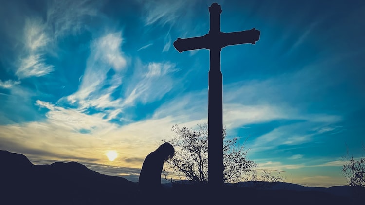 Woman kneeling before a tall cross structure while sun is setting.