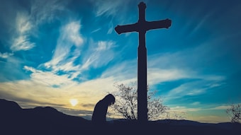 A silhouette of a person bowing in front of a large cross, with a picturesque sunset sky filled with orange, yellow, and blue hues. The scene is framed by hills in the background and sparse vegetation around the cross.