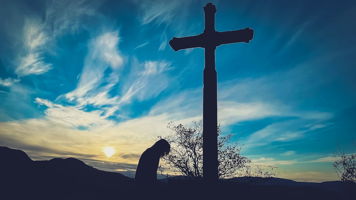 A person kneeling in prayer at the foot of a cross, silhouetted against the sky
