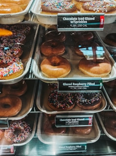 An assortment of donuts is displayed in trays behind a glass counter at a bakery. The variety includes chocolate iced glazed donuts with colorful sprinkles and plain glazed donuts. Each tray has a label indicating the type of donut and its price along with the calorie count.
