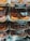 An assortment of donuts is displayed in trays behind a glass counter at a bakery. The variety includes chocolate iced glazed donuts with colorful sprinkles and plain glazed donuts. Each tray has a label indicating the type of donut and its price along with the calorie count.