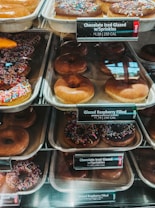 An assortment of donuts is displayed in trays behind a glass counter at a bakery. The variety includes chocolate iced glazed donuts with colorful sprinkles and plain glazed donuts. Each tray has a label indicating the type of donut and its price along with the calorie count.