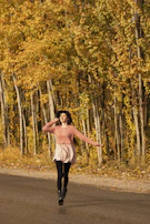 A joyful woman carrying a pastel pink Vivaluxe handbag while walking through a city street lined with autumn leaves.