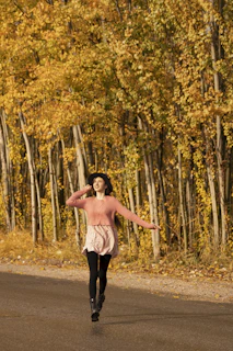 A joyful woman carrying a pastel pink Vivaluxe handbag while walking through a city street lined with autumn leaves.