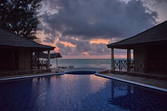 A serene beach view during sunset with two thatched-roof structures flanking an infinity pool that reflects the colors of the sky. The horizon meets the calm ocean waters, with clouds partially obscuring the warm colors of the setting sun.
