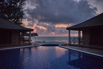 A serene beach view during sunset with two thatched-roof structures flanking an infinity pool that reflects the colors of the sky. The horizon meets the calm ocean waters, with clouds partially obscuring the warm colors of the setting sun.
