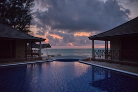 A serene beach view during sunset with two thatched-roof structures flanking an infinity pool that reflects the colors of the sky. The horizon meets the calm ocean waters, with clouds partially obscuring the warm colors of the setting sun.