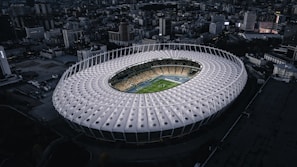 A large, modern circular stadium is surrounded by an urban cityscape. The stadium has a unique, white geometric roof design with circular patterns and is illuminated internally, highlighting a green football field and seating area. The surrounding area features a dense collection of buildings under a dimly lit, possibly overcast sky.