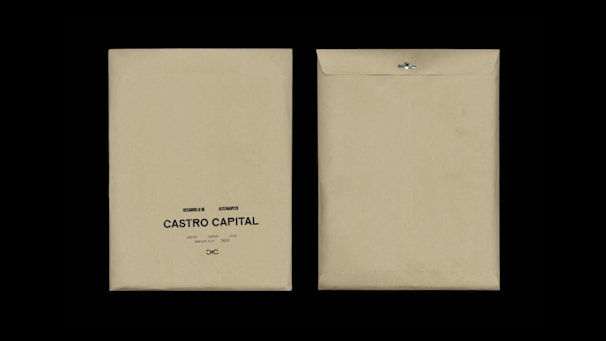 Two beige envelopes are displayed against a black background. The envelope on the left has printed text reading 'Desarrollo I, Rie, Auto Marketing, Castro Capital' with a decorative symbol below. The envelope on the right is flipped to show the plain back with a metal clasp.