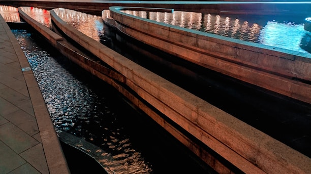 A series of curved, shallow water channels made of stone, illuminated by soft lighting. The water flows smoothly along the pathways creating gentle ripples that reflect the lights. The scene is serene and has a calming effect.
