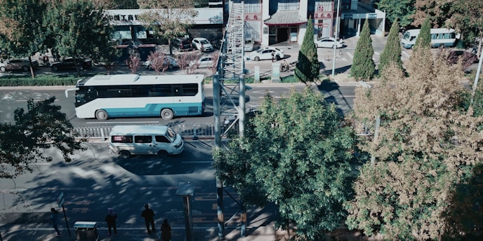An urban street view with a bus and a van traveling on the road, surrounded by trees and a few pedestrians on the sidewalk. Buildings with signage are visible in the background, under a clear sky.