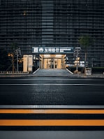 A modern building with a sleek outer facade consisting of horizontal lines and glass panels. The entrance is illuminated, displaying signage with both Korean and English text, indicating a bank. The scene includes a crosswalk with yellow safety stripes in the foreground and some greenery alongside the path leading to the entrance.