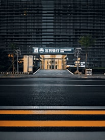 A modern building with a sleek outer facade consisting of horizontal lines and glass panels. The entrance is illuminated, displaying signage with both Korean and English text, indicating a bank. The scene includes a crosswalk with yellow safety stripes in the foreground and some greenery alongside the path leading to the entrance.