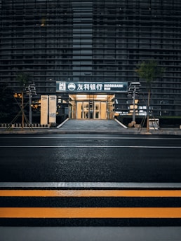 A modern building with a sleek outer facade consisting of horizontal lines and glass panels. The entrance is illuminated, displaying signage with both Korean and English text, indicating a bank. The scene includes a crosswalk with yellow safety stripes in the foreground and some greenery alongside the path leading to the entrance.