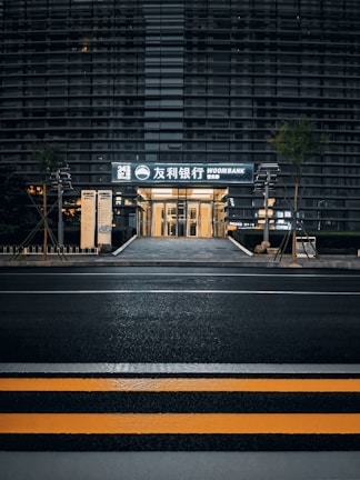 A modern building with a sleek outer facade consisting of horizontal lines and glass panels. The entrance is illuminated, displaying signage with both Korean and English text, indicating a bank. The scene includes a crosswalk with yellow safety stripes in the foreground and some greenery alongside the path leading to the entrance.
